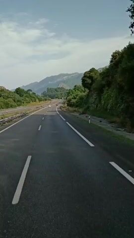 The video shows a view from a moving vehicle on a highway. The highway has multiple lanes, separated by white lines. On the left side of the highway, there is a concrete barrier and some vegetation. On the right side, there are guardrails and more dense vegetation, leading up to a hillside with trees. In the distance, mountains are visible under a partly cloudy sky. There are other vehicles on the highway, appearing small in the distance. The road is paved with asphalt.