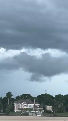 This is a vertical video shot from a moving vehicle on a highway in Fort Myers, Florida. The video captures a cloudy, overcast sky with dark, heavy storm clouds dominating the upper portion of the frame. Below the clouds, a line of lush green trees, including palm trees, stretches across the horizon, suggesting a tropical or subtropical climate.

Several houses are visible nestled amongst the trees. One house on the left is pink with white trim and appears to have a dock or waterfront access. Another, larger white house with a dark roof is more centrally located. The houses suggest a residential area, possibly a waterfront community.

The highway itself is visible in the foreground with a concrete barrier. Several cars are driving on the highway, indicating ongoing traffic and activity. Streetlights with a distinctive V-shaped design are also visible along the highway.

The overall mood is somber and dramatic due to the imposing storm clouds. The weather appears to be pre-storm or during a storm, contributing to a sense of anticipation or unease. The time of day is difficult to ascertain precisely due to the cloud cover, but it appears to be daytime. There is no visible text in the video. The activity is a car journey through a suburban or semi-rural area of Fort Myers under threatening weather conditions.