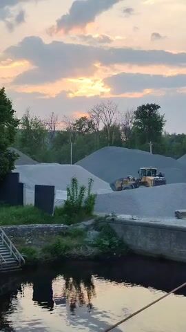 The video shows a construction site at dusk, with a yellow loader positioned on a pile of gravel. Several other piles of gravel can be seen in the background, along with trees and a partly cloudy sunset. In the foreground is a body of water with reflections of the sky and nearby structures. A set of stairs leads down to the water's edge.