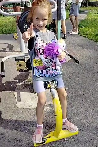 A young girl with blonde hair, wearing a grey t-shirt with a graphic print and white shorts, stands on a yellow scooter. She is holding a purple plush toy. The girl is positioned in front of an outdoor exercise machine with red and yellow components. The ground is paved with asphalt. In the background, there are trees, a fence, and other people. The lighting suggests it is daytime, likely sunny. The girl appears to be smiling.
