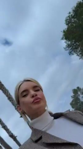 The video shows a woman standing outside with palm trees visible in the background. The sky is overcast. The woman has blonde hair pulled back in a ponytail, is wearing a white turtleneck and a brown plaid blazer. She looks up and purses her lips.