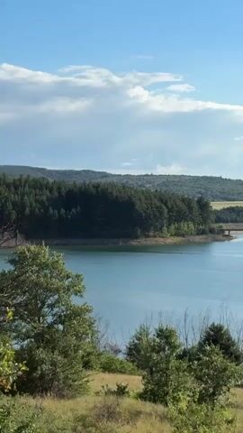 A wide, still body of water is visible under a bright blue sky dotted with white clouds. The water is bordered by a dense, dark green forest on the left bank. In the distance, rolling hills covered in greenery can be seen, with some fields of lighter green and brown interspersed. A bridge spans across the water in the middle ground, connecting the two sides. A small structure sits on a small island in the middle of the water, closer to the right side. In the far distance, on the right side, several wind turbines are visible on the horizon. The foreground is filled with dry, golden grass and scattered green trees and bushes. The overall scene depicts a natural landscape on a clear day.