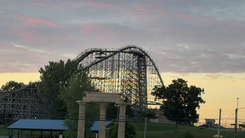 The video is a static shot of a wooden roller coaster at an amusement park. The roller coaster is the main subject and takes up a significant portion of the frame. The structure of the roller coaster, with its intricate wooden supports and track, is clearly visible. The sky above is filled with clouds, some of which are tinged with pink and orange, suggesting it might be sunrise or sunset. In the foreground, there are trees and some architectural elements that appear to be ruins or decorative structures. There are no people visible in the frame and no actions are taking place. The overall scene is calm and stationary.