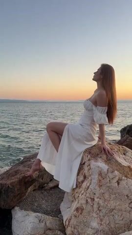 A woman with long auburn hair sits on large rocks by the sea. She wears a white off-the-shoulder dress with ruffled sleeves and a high slit on the left side. Her left leg is bent, and her right leg is extended. She is looking up and to the right with her right hand touching her hair. The sea is to her left, and the sky is clear with pastel colors of sunset. The rocks are various shades of brown and white.