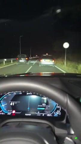 The video shows a first-person view from inside a car driving at night on a road. The dashboard displays the car's speed, which fluctuates between 42 and 48 km/h, the time as 0:47, and other vehicle information such as battery percentage and outside temperature of 25 degrees Celsius. The road is dark with streetlights and the headlights of other cars visible ahead. Two cars are in front of the vehicle, one in the left lane and another in the right lane. A triangular warning sign is visible on the right side of the road. In one brief shot, a side view of the car shows passengers inside.