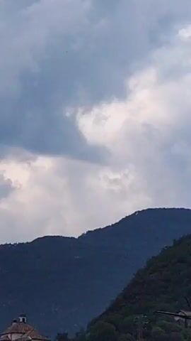 This media file shows a view of rolling, tree-covered mountains under a cloudy sky. In the foreground, a small, light-colored building with a domed roof, possibly a church or chapel, is visible on the left. The mountains are dark blue-green, with their tops silhouetted against the lighter clouds. One mountain range stretches across the middle of the frame, while another, steeper slope is visible on the right. The sky is filled with dramatic, gray and white clouds, suggesting overcast or potentially stormy weather. There are no people visible in the frame. The scene appears to be a rural or mountainous landscape, likely in the late afternoon or evening given the diffused light and cloud cover. The location is identified as Bolzano, Italy.