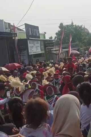 A crowded street scene is depicted, with a parade or procession in progress. In the foreground, the backs of several people are visible, including a child being held by an adult. The middle ground is filled with a marching band dressed in colorful uniforms, featuring red, green, and white, with tall, feathered headdresses. Some band members appear to be playing instruments, possibly drums. The crowd is dense, with many spectators lining the street. Indonesian flags are visible on poles throughout the scene, suggesting a nationalistic event or celebration. The background shows buildings and a dome-shaped structure, possibly a mosque, under a bright, sunny sky. The overall atmosphere appears festive.