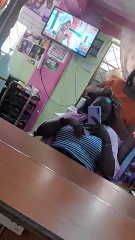 Inside a hair salon, a seated woman wearing a striped shirt and a pink shawl takes a selfie with her purple smartphone. Another woman, wearing an orange top, stands behind her and tends to her hair. The salon walls are decorated with posters of hairstyles and beauty products. A television screen shows a video. A cart with hair products is visible in the background. The scene is reflected in a large mirror in front of the seated woman.
