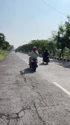The video shows a paved road in Siring, Indonesia, on a clear, sunny day. The road has some cracks and wear. There are trees lining both sides of the road, with a grassy area to the left and a landscaped area with more trees to the right. The sky is bright blue and clear, suggesting it is daytime.

The primary subjects are people riding motorcycles. In the foreground, a person wearing a pink helmet and a white outfit is riding a scooter away from the camera. Further down the road, another person on a dark-colored scooter is also riding away. In the distance, several other motorcycles can be seen on the road, indicating traffic.

The activity taking place is travel along the road. The atmosphere appears peaceful and ordinary, with no indication of urgency or distress. The lighting suggests the sun is high in the sky, likely midday. The presence of a power pole on the left side of the road indicates it's a developed area. The lush green trees and the style of the road suggest a tropical or subtropical climate. There is no visible text in the video.