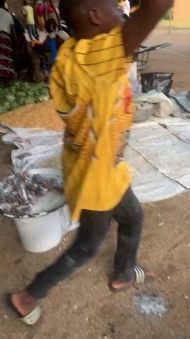 The video opens with a view of a marketplace with people and produce. In the foreground, a white bucket holds a pile of small, dark, wrapped items, possibly food. To the right, a white tarp is spread on the ground with two large mounds of grain, one yellow and one lighter beige. Behind this, a pile of green leafy vegetables, likely cabbage, is visible. Several people are in the background, some appearing to be vendors or shoppers. A woman in a yellow and black patterned dress is holding a baby. Another woman in a blue and white floral dress is walking away from the camera, holding a piece of cardboard. The environment appears to be outdoors with sandy ground and some foliage. The lighting suggests it is daytime. The video is shaky and appears to be handheld.