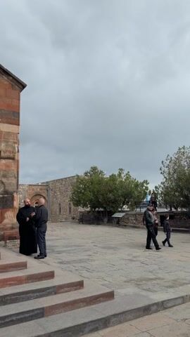 Outside a stone and brick church, a monk in black converses with a man in black jacket and pants. The two stand on the church's steps. After a moment, the monk goes inside. The exterior of the church features stone archways and steps. A stone wall is visible in the background, along with some greenery and a cloudy sky. People walk by the scene.