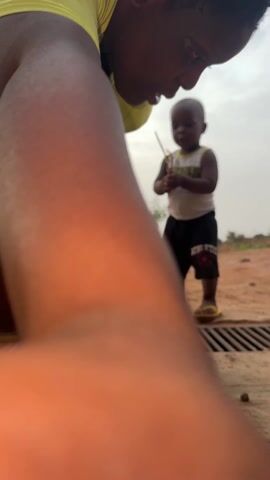 A young Black woman and a toddler are seen outdoors next to a storm drain. The woman, wearing a lime green shirt and black shorts, dances playfully in front of the toddler, who is dressed in a tank top and athletic shorts. The toddler holds a small stick in his hand. The background features an overcast sky and sparse vegetation.