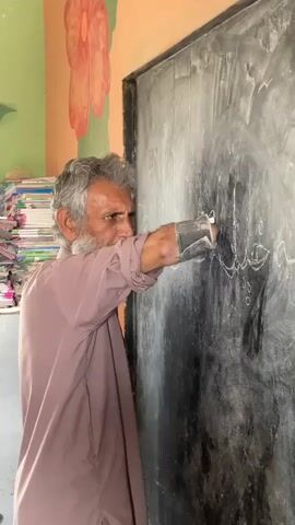 In a medium shot, an older man with gray hair and a beard is writing on a black chalkboard with white chalk. He is wearing a long-sleeved pink shirt and appears to be using a prosthetic or some kind of support on his writing arm. The writing appears to be in Urdu. Behind the man to the left is a stack of papers and books. The wall is painted with a large flower design. The scene is brightly lit, suggesting an indoor setting, possibly a classroom.