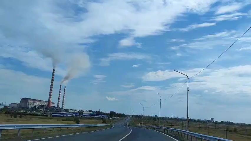 This is a first-person perspective video from inside a moving vehicle. The camera is looking forward, showing a two-lane road curving gently to the right. On the left side of the road, there is a power plant with three tall, red and white striped smokestacks emitting white smoke into a blue sky with white clouds. There are also other buildings associated with the power plant. On the right side of the road, there are several streetlights and a fence. The landscape appears to be dry and grassy. The video is only a few seconds long, and no people are visible.