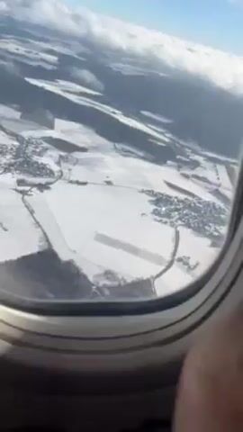 The video shows a view of snow-covered landscape from an airplane window. Patches of houses and fields are seen interspersed with roads. Some hills are visible in the background. A partly cloudy sky is visible above.