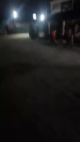 The video shows a dark street at night. A vehicle with bright headlights is seen in the distance. The camera appears to be moving, possibly held by someone walking or in another vehicle. The street is dimly lit by streetlights and the vehicle's headlights. There are buildings or structures visible in the background, but they are hard to discern in the darkness. A brief burst of sound like a horn can be heard.