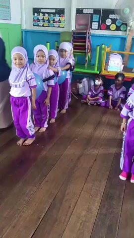 A group of young children, dressed in purple and white hijabs and outfits, are lined up in a classroom. They are holding blue balloons between their knees as part of a game or activity. The children appear to be kindergarten or preschool age. The classroom has wooden floors and colorful decorations on the walls. Some other children are sitting on the floor in the background. The overall atmosphere seems playful and educational.
