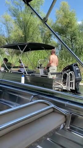 The video captures a serene moment on a calm body of water, likely a river or a lake, surrounded by lush greenery and trees. In the foreground, the interior of a metal boat is visible, with comfortable-looking cushioned seats and chrome railings, suggesting a leisurely excursion. The camera appears to be positioned from within this boat, looking towards another boat ahead.

The second boat, also metal, is powered and moving. It has a canopy providing shade, and three people are on board. The person at the helm, a man with his back to the camera, is steering. Two other individuals are seated in the boat, one facing forward and another slightly turned. They appear relaxed, enjoying the ride. The boat has a white buoy attached to its side, and a visible website address is printed on its hull: "www.alunaetika.rs".

The surrounding environment is abundant with tall, green reeds and aquatic plants, indicating a natural, perhaps marshy, habitat. The trees in the background are full of dense foliage, suggesting it is a warm season, likely summer. The sky is a clear, bright blue, hinting at a pleasant, sunny day. The overall atmosphere is tranquil and peaceful, evoking a sense of calm and enjoyment of nature. The time of day appears to be daytime, given the brightness of the sunlight. There are no obvious signs of any specific event, but rather a scenic boat trip. The location is identified as Prevlaka, Montenegro.