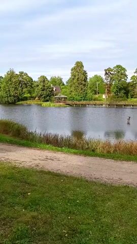 The video pans across a serene lake surrounded by lush green trees. A wooden pier extends into the water, and a gazebo is visible on the shore. The sky is overcast with a few clouds. In the foreground, a dirt path runs alongside a grassy area.