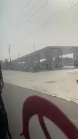 The video is shot from inside a moving vehicle, showing a view of a street with a gas station on the right. The weather appears hazy or dusty. A few people are walking near the gas station, carrying bags. The road is wet. The camera angle is slightly obstructed by the car's interior.