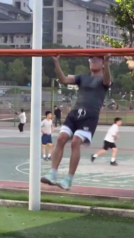 The short video clip captures an outdoor fitness area, likely in a park or university campus in Jiangjin, China.

In the foreground, a man wearing a dark gray t-shirt, black athletic shorts with white undershorts, light blue-green athletic shoes, and a dark baseball cap performs one pull-up on a red horizontal bar. He executes the pull-up with a controlled motion, pulling himself up until his chin is above the bar, then lowering himself back down. After completing the exercise, he steps away from the bar towards the left side of the frame.

As the first man moves away, a second man, dressed in a black t-shirt and light gray pants, with white wireless earbuds, enters the frame from the right. He raises his arms slightly, appearing to be stretching or preparing to use the pull-up bar himself.

The background of the scene features a green grassy area adjacent to a marked basketball court with red and green sections, and a basketball hoop visible in the distance. Several other individuals, including adults and a young boy in a yellow and blue shirt, are scattered around the court area, engaged in casual activities or walking. Beyond the sports area, multi-story residential or dormitory buildings with light-colored facades and dark roofs dominate the far background.

The lighting suggests it is late afternoon or early evening, with soft, ambient daylight indicating clear weather. No specific text is clearly legible on any clothing or signs. The overall atmosphere is tranquil and active, with people enjoying outdoor exercise and leisure.