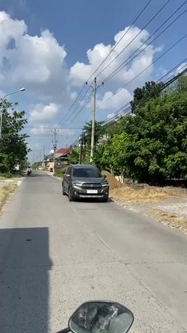 The video is a first-person perspective of someone riding a motorcycle down a paved road on a sunny day with a blue sky and scattered clouds. On the left side of the road, there are trees, a street lamp, and some buildings in the distance. On the right side, there are more trees, power lines running along utility poles, and several buildings, some with covered awnings and others appearing to be houses. A gray SUV is parked on the right side of the road in the foreground. Further down the road, a motorcycle with a rider is visible. The road surface appears somewhat worn with some cracks. Shadows from trees are cast across the road, indicating the time of day is likely late morning or early afternoon. The overall environment appears to be a semi-urban or suburban area.