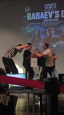 A group of men are gathered around an arm wrestling table. One man, wearing a black and white striped shirt, acts as a referee and is holding the hands of two competitors. Another man, wearing a white shirt, stands to the right of the table, seemingly observing the match. The competitors are positioned on opposite sides of the table, gripping each other's hands in a starting position for arm wrestling. A screen is visible in the background, displaying text that includes "KING OF TABLE PRESENTS - BABAEV'S CUP". The event appears to be taking place indoors in a room with a wooden ceiling. Red rope barriers are set up in the foreground, separating the audience area from the competition space.