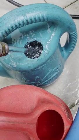 The video shows a close-up view of two watering cans, one teal and one red, on a textured surface that appears to be concrete or stone. A metal nozzle from a hose is being inserted into a large, irregularly shaped hole in the top of the teal watering can. Water is flowing forcefully from the nozzle, creating a splash and filling the teal watering can, some of which is overflowing onto the surface. A hand, with visible fingers and thumb, is holding the hose nozzle in place. A blue hose is also visible, extending from the nozzle. The red watering can is positioned in the foreground, with its opening visible. The overall scene suggests an outdoor setting, possibly a garden or patio, with natural light. No text is visible in the video. The activity is focused on filling the teal watering can, which appears to have a damaged opening. The precise time of day or weather conditions are not discernible. The location context provided is West Vancouver, Canada.