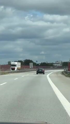 A black Audi A4 drives on a highway under a cloudy sky. The car is in the right lane, heading away from the camera. The highway has multiple lanes with white lane markings. On the left side of the highway, there is a red barrier. In the distance, there are trees, industrial silos, and buildings. The camera is positioned from inside another vehicle traveling on the same highway, capturing the Audi from behind.