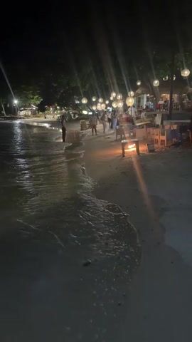 A long, high-angle shot captured from an empty, sandy beach shows gentle waves lapping at the shore on the left side of the frame while people walk and sit along the shore on the right side of the frame at night. Some of the people standing appear to be talking. There are several tables and chairs on the sand where people sit. Round, yellow lights hang from unseen structures that create light streaks in the air from their brightness. The lighting on the beach in general is dim due to the nighttime.