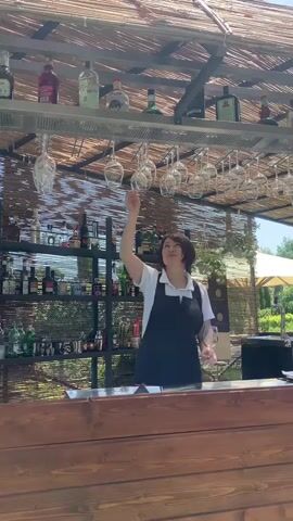In this short video clip, a woman is seen behind a wooden bar, preparing to serve drinks. She is wearing an apron and a white shirt. The bar is situated outdoors, under a thatched roof, suggesting a warm, sunny day. Shelves behind the bar are stocked with various bottles of alcohol and glassware, including wine glasses hanging from a rack above. The woman reaches up to take a wine glass from the rack, then brings it down to inspect it. She smiles at the camera, appearing friendly and professional. The setting appears to be a resort or outdoor dining area, with lush greenery visible in the background, hinting at a beautiful, possibly rural, Italian landscape. The overall impression is one of relaxed service in a pleasant, open-air environment.