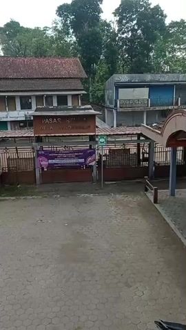 A wide shot shows a market entrance with buildings behind it. A sign overhead reads "PASAR HEWAN DESA RANDULAWANG". A banner hangs on a fence in front of the market entrance, displaying text and images. The ground in front of the entrance is paved with interlocking concrete blocks. Trees and vegetation are visible in the background.