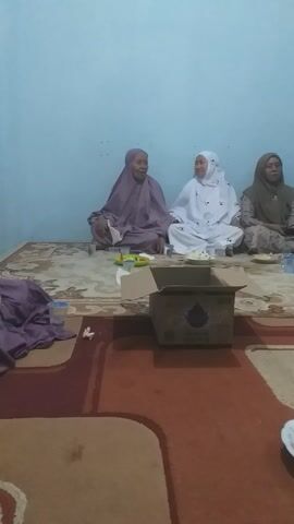 The media file captures a gathering of people, likely women, in an indoor setting. The subjects are seated on the floor on a patterned rug, facing a light blue wall. Three women are prominently visible in the foreground, all wearing hijabs. One woman in a light purple hijab is seated on the far left, looking towards the center. Next to her, a woman in a white hijab is laughing heartily, her head tilted back. To her right, another woman in a brown and beige patterned hijab is looking to her left.

In the background, through an open doorway, more people can be seen, suggesting a larger gathering. The doorway reveals a dimly lit outdoor area, possibly a patio or street, with a wooden door frame. The room itself appears to be a living space, with a patterned rug on the floor. A cardboard box is placed in front of the seated women, and plates with food items, including what looks like fruit and pastries, are also visible on the rug.

The overall mood appears to be cheerful and social, indicated by the laughter of the woman in white and the relaxed postures of the others. The lighting suggests it might be evening or nighttime. The setting and attire of the individuals point towards a cultural or religious gathering, possibly a family event or a community meeting. The presence of food suggests a meal or a refreshment break during the event. There is no visible text in the media file.