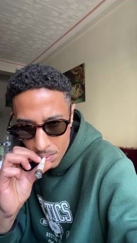 The video shows a man with curly dark hair, sunglasses, and a green hoodie smoking a joint. He exhales a large cloud of smoke towards the camera. A bottle and a painting are visible in the background. There is music playing.