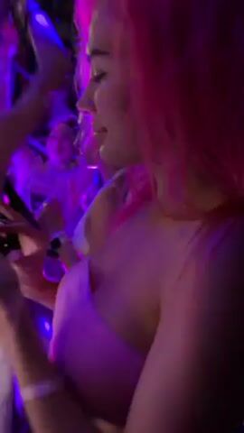 The video shows a woman with bright pink hair dancing in a crowd. She is wearing a light pink strapless top and is holding a mobile phone in her hand. The environment is brightly lit with blue and purple party lights. There are other people dancing in the background.