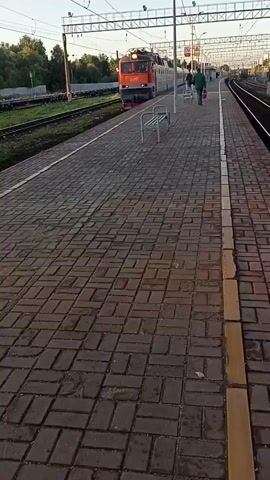A train is slowly moving past a tiled train station platform. The train is red and silver with multiple cars, and it is moving from left to right in the frame. There is an electric locomotive at the front of the train, which is orange and red. A person in a green uniform is walking on the platform in the same direction as the train. There are benches on the platform. The platform is paved with red brick. There are power lines and poles visible above the train and platform. The sky is clear and the sun is shining.