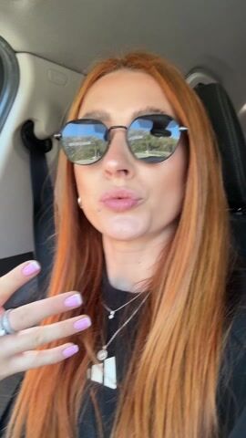 A woman with long red hair is in a car, wearing sunglasses and a black shirt. She makes a rock-and-roll hand gesture, sticks out her tongue, and runs her fingers through her hair while looking at the camera. The car's interior is visible, including the seatbelt and part of the dashboard.