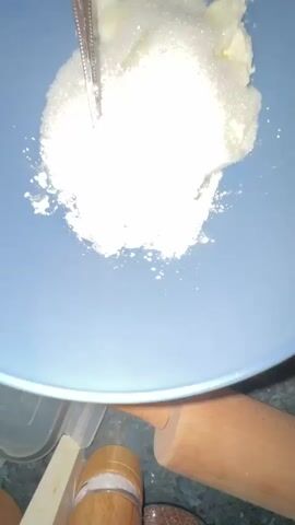 A close-up, overhead shot shows a hand using a silver fork to mix ingredients in a light blue bowl. The ingredients include what appears to be butter, sugar, and flour. The hand is fair-skinned and moves to combine the ingredients. Some of the dry ingredients scatter slightly from the mixing action. A small portion of a patterned countertop, a wooden object, and a yellow utensil are visible in the bottom of the frame.