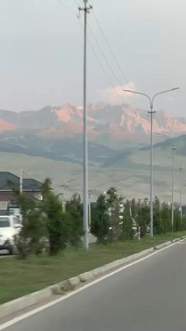 The video shows a roadside view with mountains in the distance. A paved road with a white line marking the edge is in the foreground, with a grassy verge and trees along the side. Several streetlights are visible along the road. There are a few cars on the road, moving away from the camera. In the middle ground, there are some houses and more trees. The mountains in the background are rugged and appear to be catching the light, possibly during sunrise or sunset, with some snow visible on their peaks. The sky is overcast.