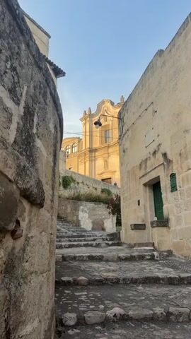 The video shows a narrow, stone alleyway in an old town. The buildings on either side are made of light-colored stone and appear ancient. A set of stone steps leads upwards into the alley. At the top of the steps, a building with a yellow facade and a large dome can be seen. The sky is clear and blue. The time of day appears to be either early morning or late afternoon, as the sun is casting a warm glow on the buildings. There are no people visible in the video. The overall impression is one of historical charm and tranquility.