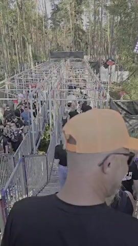 The video shows a group of people walking up a metal walkway constructed with scaffolding. The walkway appears to be elevated and is surrounded by trees. There are lights and sound equipment visible, suggesting an outdoor event or festival. People are dressed casually, and some are carrying backpacks. The walkway has handrails on both sides, and a black tarp is visible along one side. The ground below the walkway is covered in grass and trees. The camera is positioned behind the group, looking forward and slightly upwards. The overall atmosphere appears to be busy and energetic.