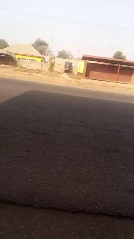 The video shows a street scene on a clear day. The camera is stationary, positioned near the edge of a paved road. In the background, there are buildings, including one painted yellow with a light gray roof, and a brown bus stop-like structure. Power lines are visible overhead. A yellow and green bus passes by on the road. The pavement is cracked and shows signs of wear. A shadow falls across the foreground.