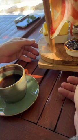 The video pans across a wooden table laden with food and drinks. A person's hands are seen adjusting a tiered wooden serving platter. The platter holds various items including slices of cured meat, cheese, olives, a dollop of dark jam, a red spread, and white cream. In the background, other people are visible, and drinks in glasses and cups are present. One person is seen holding a small terracotta pitcher. The overall setting appears to be an outdoor dining area, with sunlight casting shadows on the table.