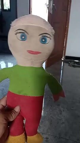 The video shows a close-up of a homemade doll being held by a hand. The doll has a light-colored fabric head with blue eyes, painted red lips, and what appears to be black hair or a black fabric patch on top. It is wearing a green shirt and red pants with yellow fabric for the feet. The background is slightly out of focus, showing a tiled floor and what looks like a door and a wall. The doll's head is blurred, suggesting it is either moving or the camera is focusing on the body.