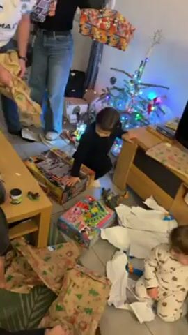 The video shows a living room during a gift-opening event, likely Christmas morning, with wrapping paper scattered around. A small Christmas tree stands near a television stand. Multiple people are visible, including adults and young children. One child in black is near several unwrapped toy boxes. Another younger child is wearing pajamas with a pattern. Adults hold wrapped gifts with cartoon-themed designs. A small wooden table holds a few small objects. The room is brightly lit, suggesting daytime.