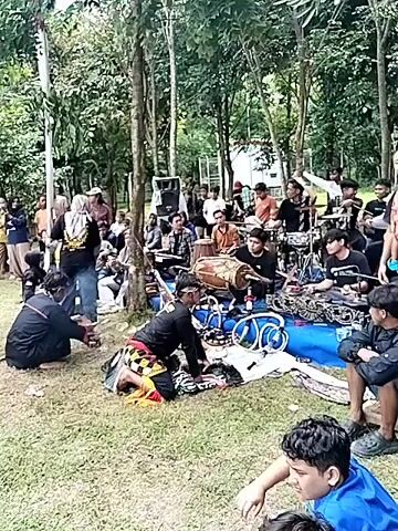 The video shows an outdoor performance on a grassy field, surrounded by trees and a crowd of spectators. Initially, the focus is on musicians playing traditional instruments and a person performing some kind of ritual or dance on the ground. The camera then zooms out to reveal a wider view of the field, showing performers in black clothing, possibly engaged in martial arts or a cultural display. In the background, there's a banner with text, indicating an event or ceremony. There are flags visible. The overall scene suggests a cultural event or public performance taking place outdoors.