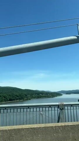 The video depicts a view from a bridge overlooking a wide body of water. The water is surrounded by lush, green, forested hills. The sky is clear and blue. The camera is positioned behind the bridge's railing and concrete barrier. A large, cylindrical pipe and cables form part of the bridge's structure in the foreground. A highway number sign "63" is visible on the railing. There are boats visible on the water.