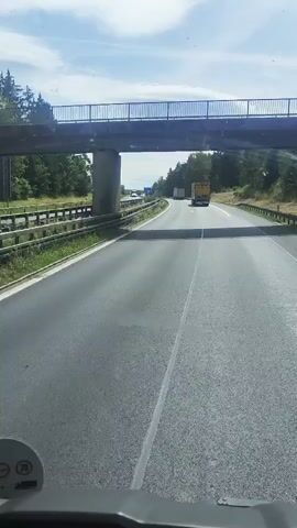 This is a first-person perspective video of someone driving on a highway. The road has multiple lanes, and there are other vehicles on the road, including trucks and cars. The highway is lined with trees and vegetation, and there is an overpass in the distance. The weather appears to be partly cloudy. The video shows the road ahead, with the dashboard of the vehicle partially visible in the foreground.
