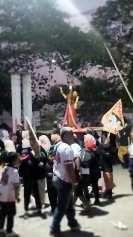 The video depicts a group of people gathered outdoors, seemingly at dusk or night, with some ambient light sources illuminating the scene. A vertical white pole with decorative elements, possibly pillars or columns, stands prominently in the background. Several individuals are visible, with some wearing white shirts and others in dark clothing. One person is holding a large, light-colored flag or banner featuring a character that appears to be Mickey Mouse. Another person is carrying a flag with red and white stripes. The crowd is positioned in front of a backdrop of trees and foliage. The overall atmosphere suggests a public gathering or event.