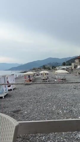 The media file shows a view of a pebbly beach in Vallecrosia al Mare, Italy. The scene is set under an overcast sky, suggesting a possibly cool or cloudy day. In the foreground, a white wicker chair and table are partially visible, implying the viewpoint is from a seated position, perhaps at a cafe or restaurant overlooking the beach.

The beach itself is lined with white cabanas, each with white curtains that are partly drawn, creating a sense of privacy and tranquility. These cabanas are set up on wooden platforms. Several beach chairs and umbrellas are also scattered across the beach, some occupied by people relaxing. A few individuals can be seen walking along the shoreline or sitting near the water.

In the background, the calm sea stretches out towards a horizon defined by rolling hills and mountains that are softly lit by the diffused daylight. The muted colors of the landscape and the cloudy sky contribute to a peaceful, somewhat subdued atmosphere. The overall impression is one of a quiet seaside resort, possibly in the late afternoon or early evening, before the main rush of beachgoers or after they have dispersed. There is no visible text in the media file.